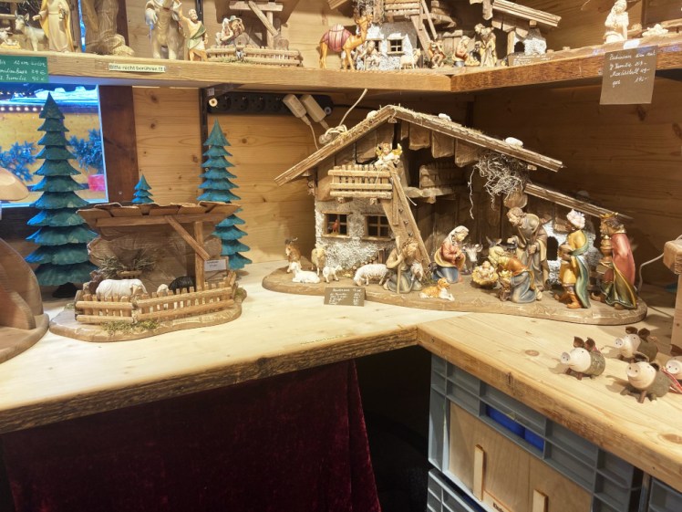 Two shelves of very elaborate handmade wooden stable sets filled with figures. To the right is a set of very cute carved slightly cartoonish pigs.