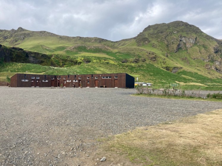 A long low dark brown building in front of a gravel patch. Behind it is a grassy mountain with a craggy top.