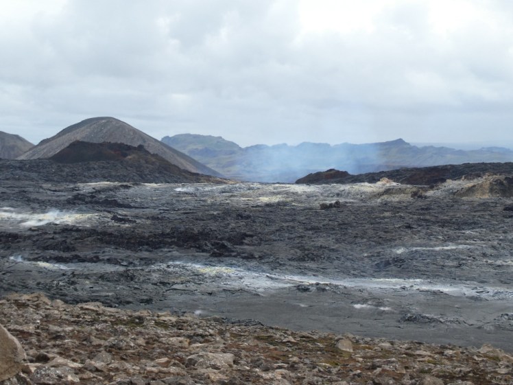 A field of lava with the texture of the top of chocolate brownies, all streaked with white sulphur deposits. A fresh dark crater is on the horizon and behind that, blue smoke rises up from the 2021 eruption.
