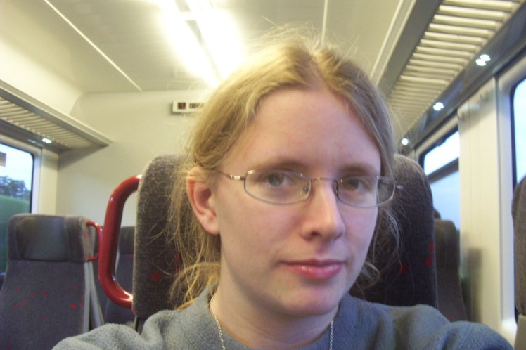 Idiot student train selfie on the way back. I'm clearly holding the camera in both hands. My hair is tied back, I'm wearing wire-framed glasses and a grey-green fleece sweatshirt.