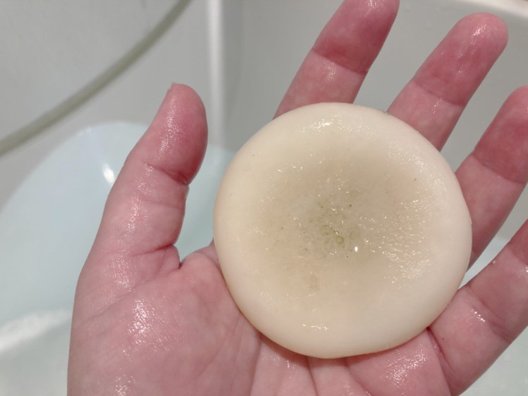 A beige-white round conditioner bar with a dent in the middle lying in my red wet hand. The bar is a bit grey-brown in the middle, which is because it's been in use for over a year.