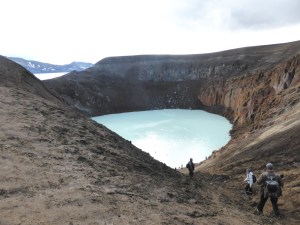 A muddy crater with a blue lake in the bottom. People are climbing down the side of the crater and there are other people, who look like dots from here, swimming in the warm water. In the background is a hint of a ring of mountains. These are the caldera edge and the glimpse of water beyond is the extremely deep and scary Askja lake, where the caldera is flooded.
