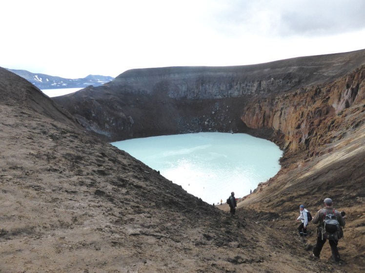 A muddy crater with a blue lake in the bottom. People are climbing down the side of the crater and there are other people, who look like dots from here, swimming in the warm water. In the background is a hint of a ring of mountains. These are the caldera edge and the glimpse of water beyond is the extremely deep and scary Askja lake, where the caldera is flooded.