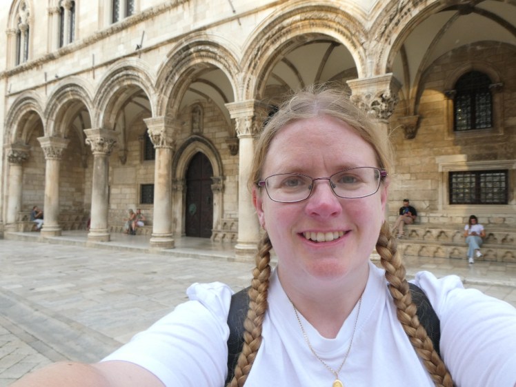A selfie (wearing a white t-shirt) in front of the rounded arches of the Rector's Palace. The stone is either golden-white or stained yellowish with age.