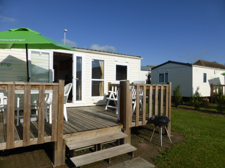 A Eurocamp static caravan in the sun. It has wooden decking outside and a big green umbrella.