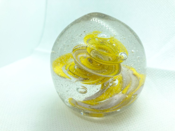 The finished paperweight, a chunk of glass nearly two inches in diameter with a tight swirl of bright yellow glass inside. You can also make out a bit of pale pink swirl.