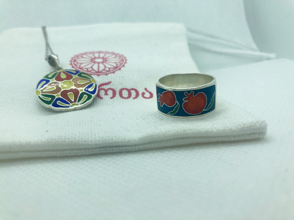 My finished pendant, a geometric floral pattern in a circle, and a ring with pomegranates on a navy background, lying on a white bag printed with the Ikorta logo.