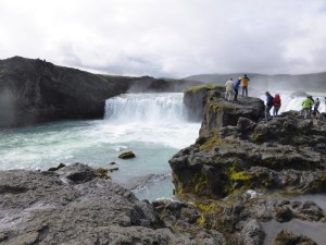 Goðafoss, a horseshoe-shaped cascade falling over fairly low black basalt cliffs. The churned water is greenish-white and the sky overhead is heavy with white clouds.