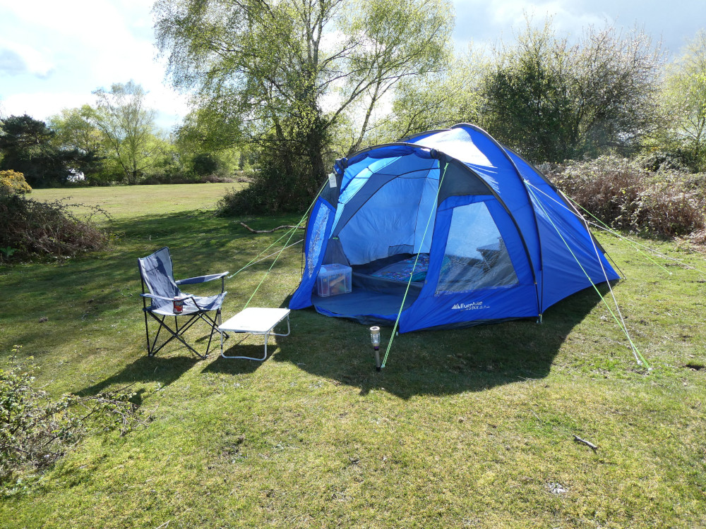 My tent, a large blue dome with a porch, open to the sun. Outside is a folding chair and tiny table. The whole setup is in a little clearing in the campsite with trees behind it.