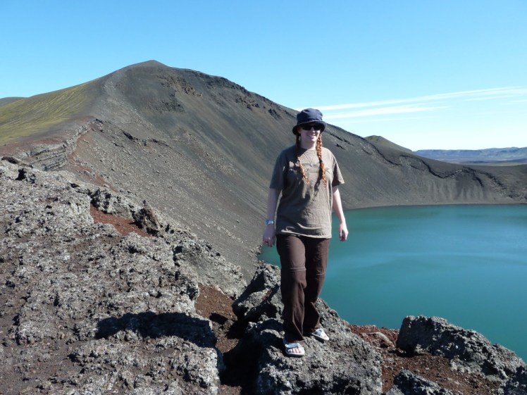 Me in a green-grey t-shirt standing on the edge of a crater with reddish rocks around the edge and flooded with bright blue water.