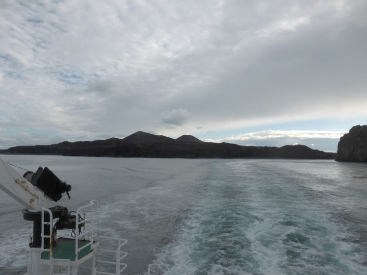 Sailing away from Heimaey. The island is silhouetted but two volcanoes are clearly visible on its skyline.