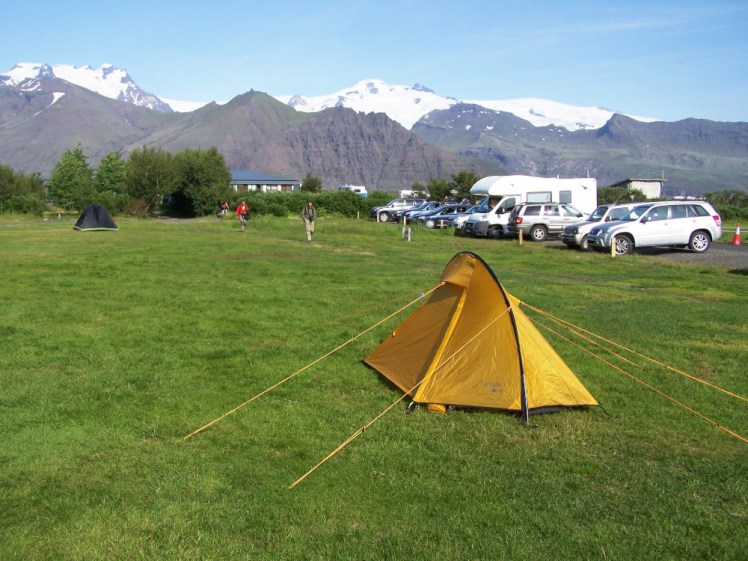 A small yellow tent pitched on some very green grass. Behind the camping field is a very bare mountain side and above that, snow-capped mountains concealing the true extent of the glacier behind.