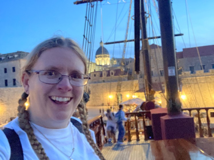 Me on the deck of a tall ship with the walls of Dubrovnik behind me. It's early evening, just dark enough that the town lights are really bright.