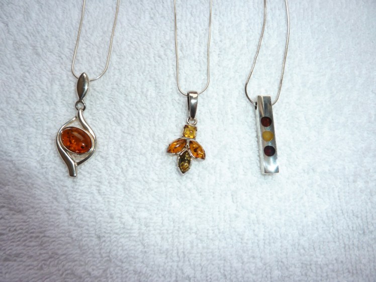 Three different styles of amber necklace on silver chains. The first is a large orange oval with a silver curved eye-shaped frame. The second is three colours in a flower or insect shape and the third is a silver bar with three circles of three different colours of amber set within.