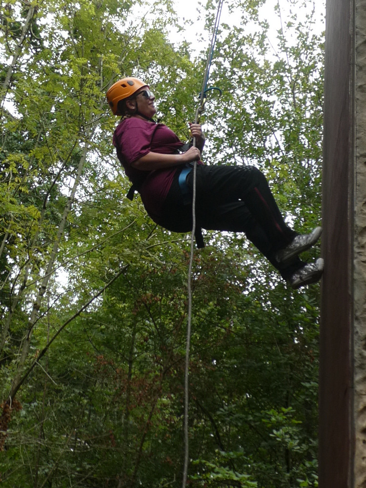 Me, in an orange helmet and sparkly trainers, hanging from a rope against an abseiling wall. Behind me are lots of trees because this is in the woods.