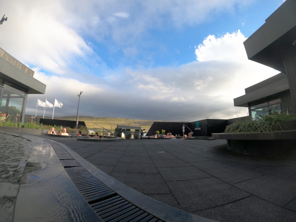 Taken from the edge of the shallower warm pot at the back: you can see the two raised hotter pots in front. Behind them are the lower pot that sticks out the front and the shallow one off to the left, which you can't really see because it's behind the relax room. You can see the glass windows and part of the square roof. Out the front, you can see the sun shining on the mountains beyond and I think you can see a hint of steam from the hot spring.