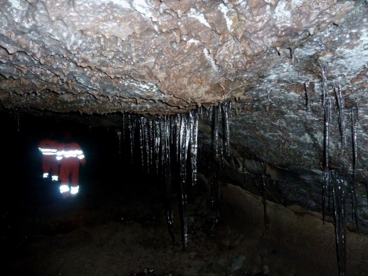 Glittering stalactites in a lava tube. The cave itself is dark and the rock is reddish. In the distance are two people wearing warm suits with reflecting stripes on the back, reflecting very brightly.