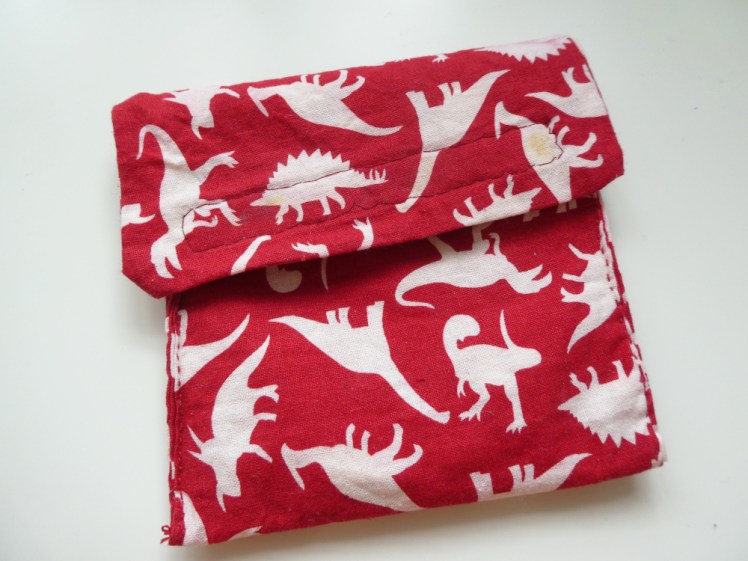 A small square pouch with a velcro flap. It's made of red fabric with white dinosaurs. The dinosaurs are a bit pink in places and there are glue stains on some of the dinosaurs from a failed attempt to glue the velcro on.