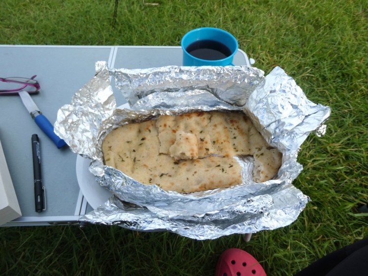 A piece of garlic bread nestled in silver foil. Behind it is a tiny camping table with an enamel mug of squash on it.