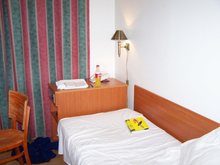 My private room in a now-closed hostel in Helsinki. There is a narrow single bed with white bedding, a table at the end, a chair visible to the side and the Finnish winter is blocked out by red and green striped curtains.