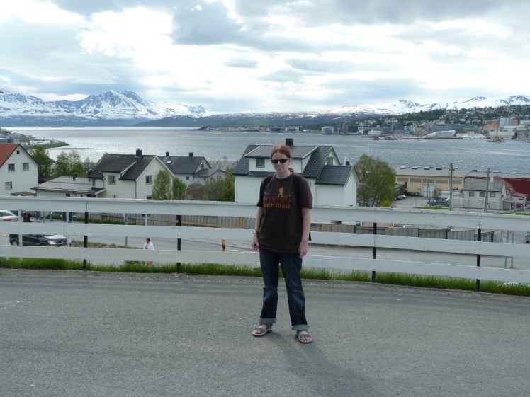 Me, in t-shirt, sandals & sunglasses in the Arctic Circle in May. Behind me, the harbour of Tromso is surrounded by snowy mountains.