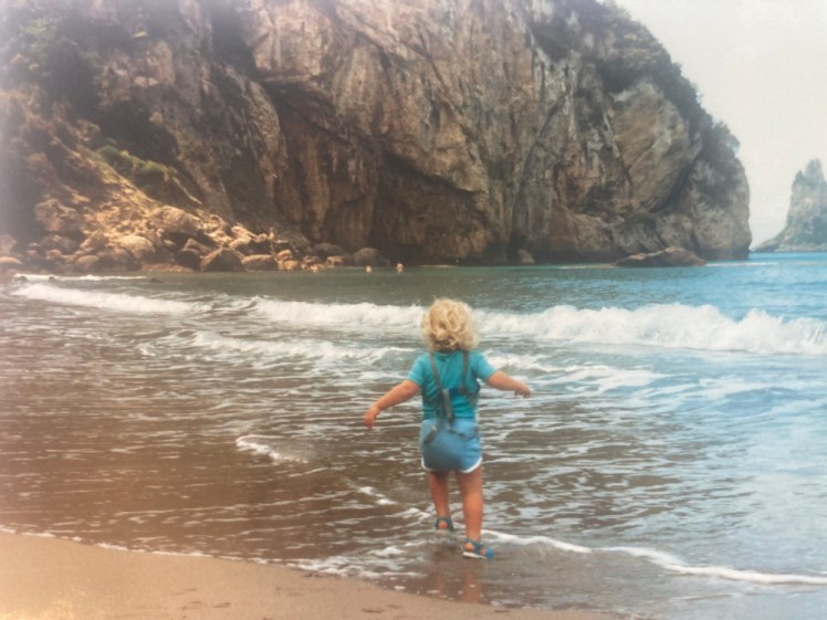 Me prancing around in the shallows in the sea, in front of some spectacular cliff formation. I'm wearing a blue t-shirt and shorts and also a set of reins.