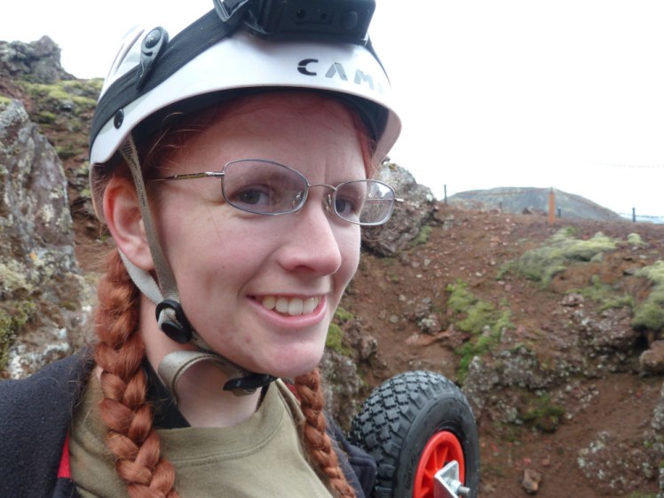 A photo, probably a selfie, from a once-in-a-lifetime day trip, inside the volcano. I'm wearing a helmet with a lamp on it, a green t-shirt, my hair is redder than usual and in plaits. My glasses are blue-tinted and pulled skew-whiff by the helmet straps.