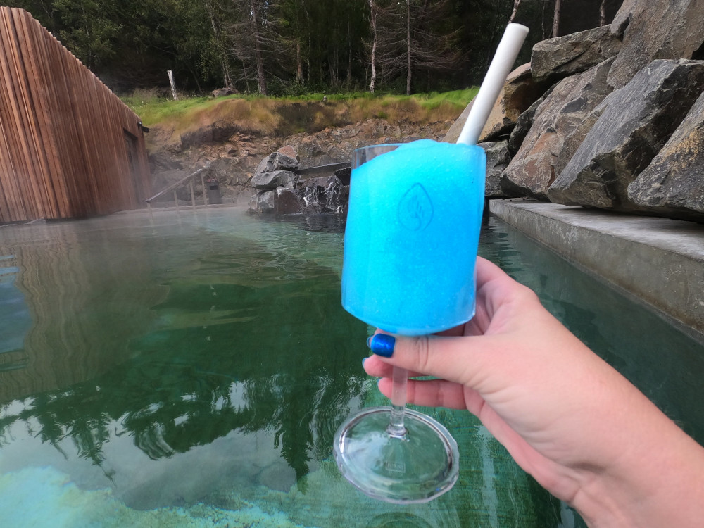 A (reusable) plastic wine glass containing a bright blue slushie. My nails are a beautiful glittery blue too and behind the drink, you can see the green water of the lagoon.