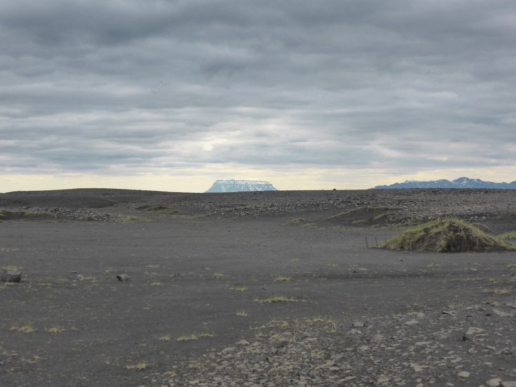 A grey featureless and utterly bleak lava field. In the distance and hazy is a flat-topped mountain standing on its own.