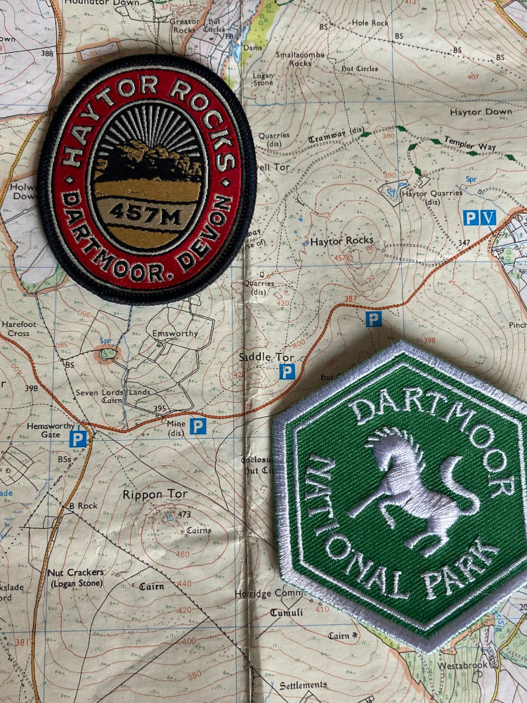 An oval Haytor badge with a red circle around the inside and a picture of the tor with sunlight radiating behind it, and a green hexagonal badge with a white horse and Dartmoor National Park on it. They're both lying on my OS map showing the area I walked today.