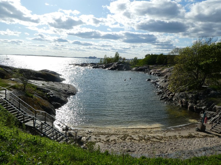 A narrow shallow inlet with rocky sides and yellow sand and foam at the end. Steps lead up one side and you can make out Helsinki on the horizon.