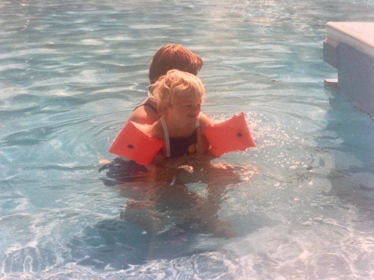 Me as a toddler, with very blonde hair, in a swimming pool, held by my mother and wearing orange inflatable armbands.