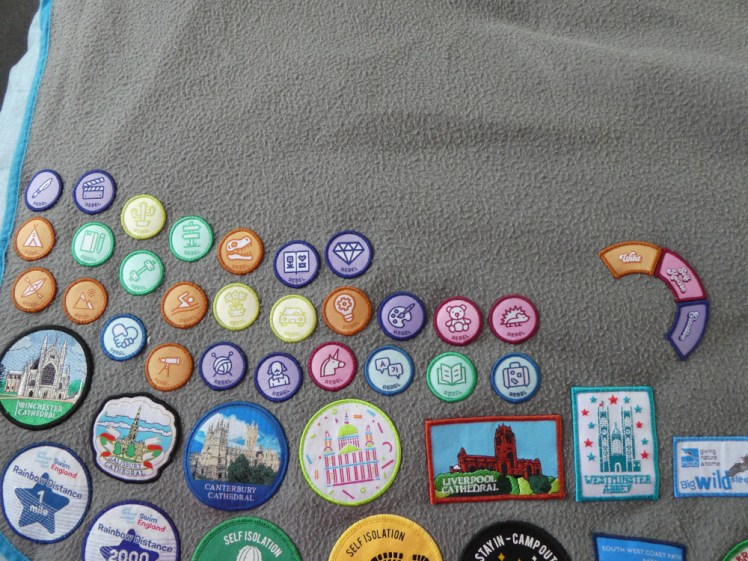 Part of my non-guiding badge collection on my grey blanket. At the top are three rows of small round Rebel badges. Underneath is a line of cathedrals and at the bottom are some swimming badges and some "life continued in 2020" badges.