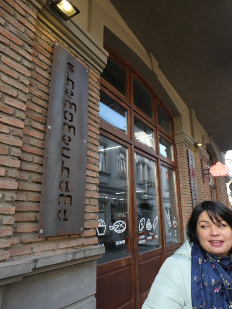 Tour guide Lela standing outside a restaurant with a steel cut-out sign with the name Shemomechama vertically down it.