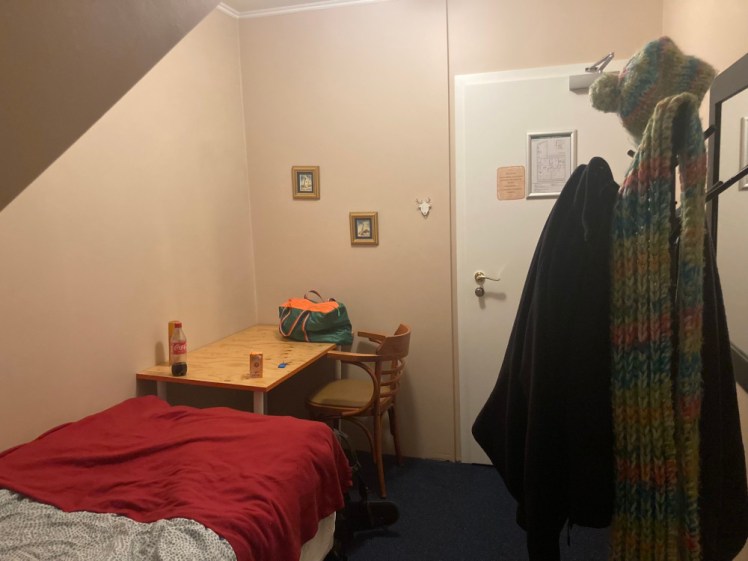 My guesthouse room in Reykjavik from February. The ceiling slopes down steeply above a single bed, I have a bag and some drinks on a wooden table and the only clothes storage is a hat stand draped in hats, scarves and jackets.
