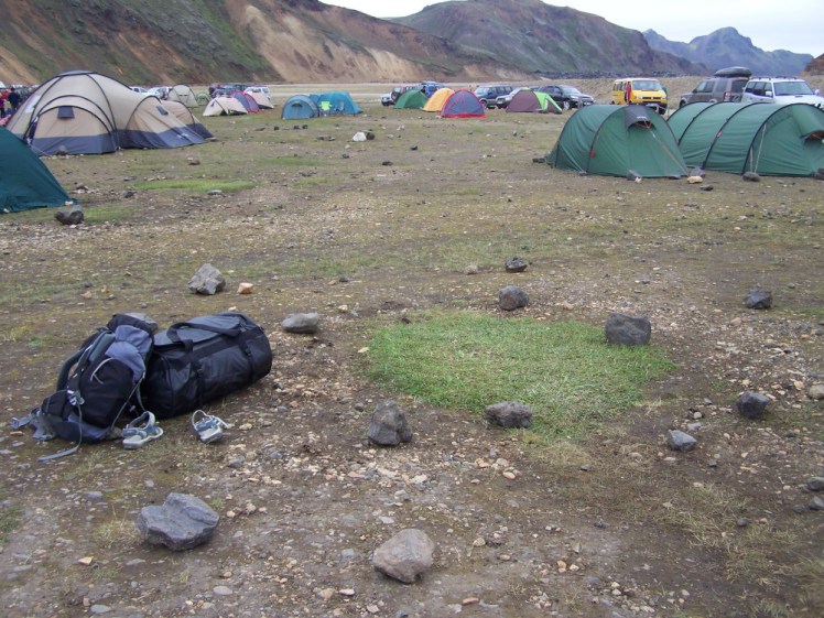 A pile of luggage - a large black duffle bag, a grey 45l backpack and a pair of sandals - lying next to a patch of grass on the gravelly stony Landmannalaugar campsite.