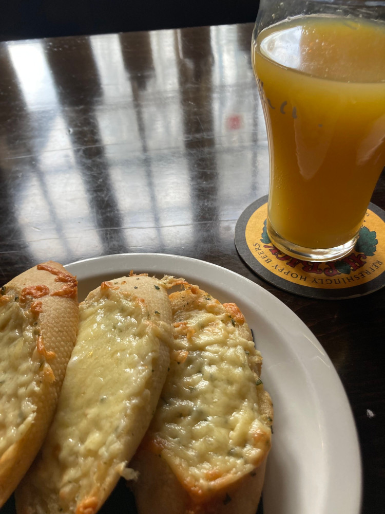 Lunch at the Millburys - three slices of cheesy garlic bread and a glass of orange juice & lemonade on a very shiny brown wooden pub table that's reflecting the window above.