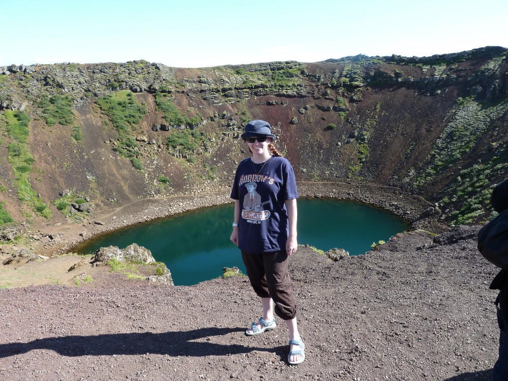 Me at Kerið crater, a small crater with steep sides, reddish rock streaked with green and a blue-green lake in the middle. I'm wearing a blue bucket hat, a blue Boardwalk Empire t-shirt and brown hiking trousers rolled up because it was an unusually hot day in Iceland.