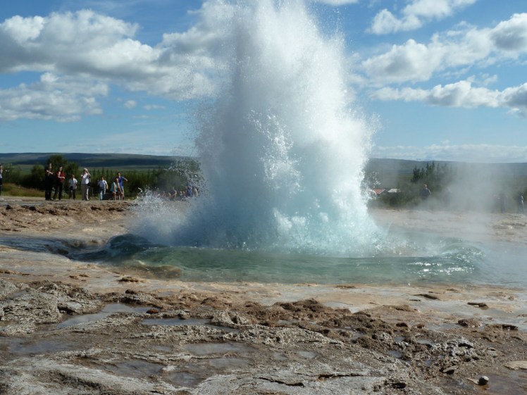 Strokkur, an active geyser, erupting. The photo has caught the very beginning of the eruption, when it's still low and wide and hasn't yet reached its peak, with blue water flying out from the base as the water blasts through the bubble that forms in the second before eruption.