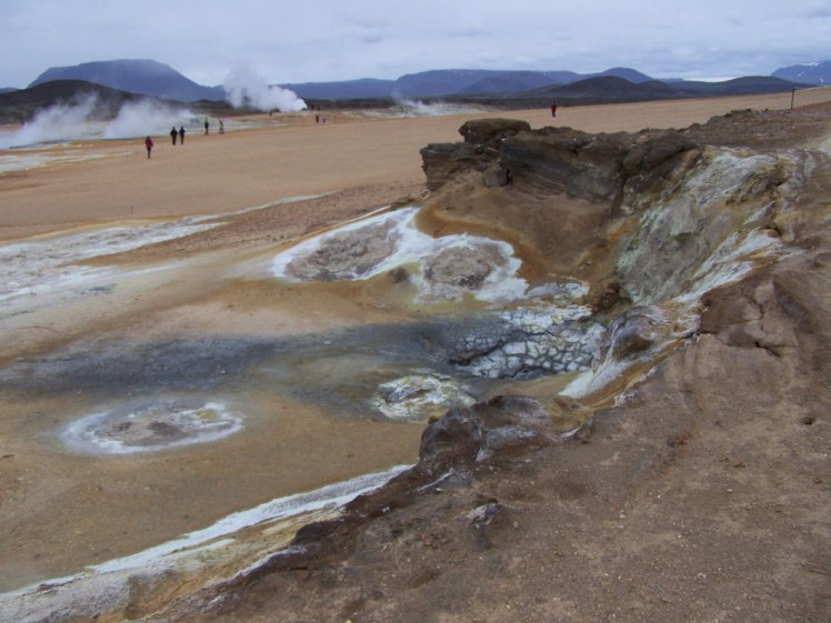 Hverir hot springs area: a flat orange sandy area at the foot of the mountains. It's streaked with patches of white and yellow and clouds of steam. This photo is taken from a miniature cliff which is equally sulphur-streaked.