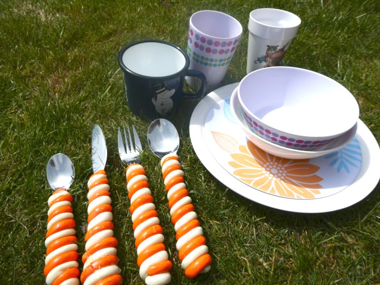 Two melamine plates, bowls and tumblers, an enamel mug and a cutlery set with orange and white striped polymer clay handles, all lying on the grass in the sun.