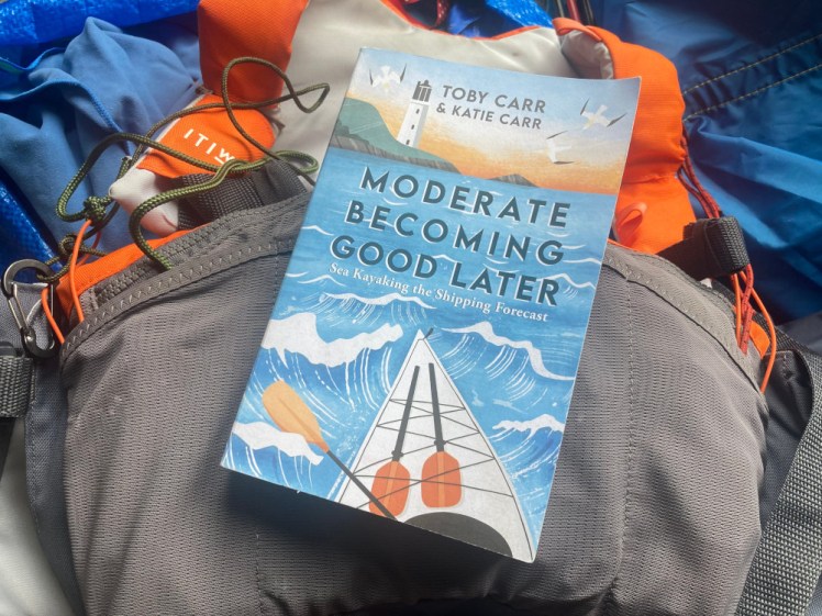 Moderate Becoming Good Later, a book with a picture of a white kayak surging out into the sea towards a lighthouse. The book is lying on my buoyancy aid.