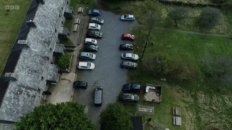 An aerial shot showing a row of grey terraced cottages on the left and a car park with far more cars than usual in it.