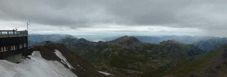 A panorama with the skyline walk to the left and a desolate grey and green mountain scene to the right. Behind, you can make out one of the lakes at Interlaken.