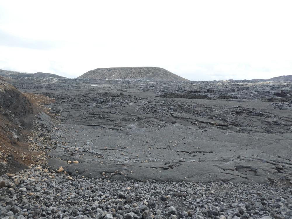 A dark greyish-brown lava field with a flat-topped crater in the background. The lava kind of has the texture of the top of a pan of brownies.