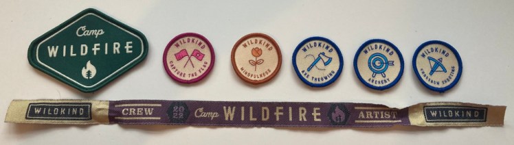 A green diamond-shaped Camp Wildfire badge followed by five round badges with coloured borders, icon representing the activity and the name of the activity. Underneath is my cream and purple festival wristband.