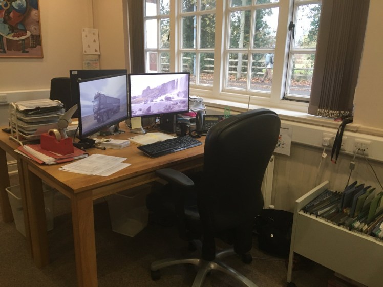 My desk at my office at work. It's a wooden kitchen table with a big comfy leather wheely chair. There are two reasonably large monitors on arms, both purple-tinted