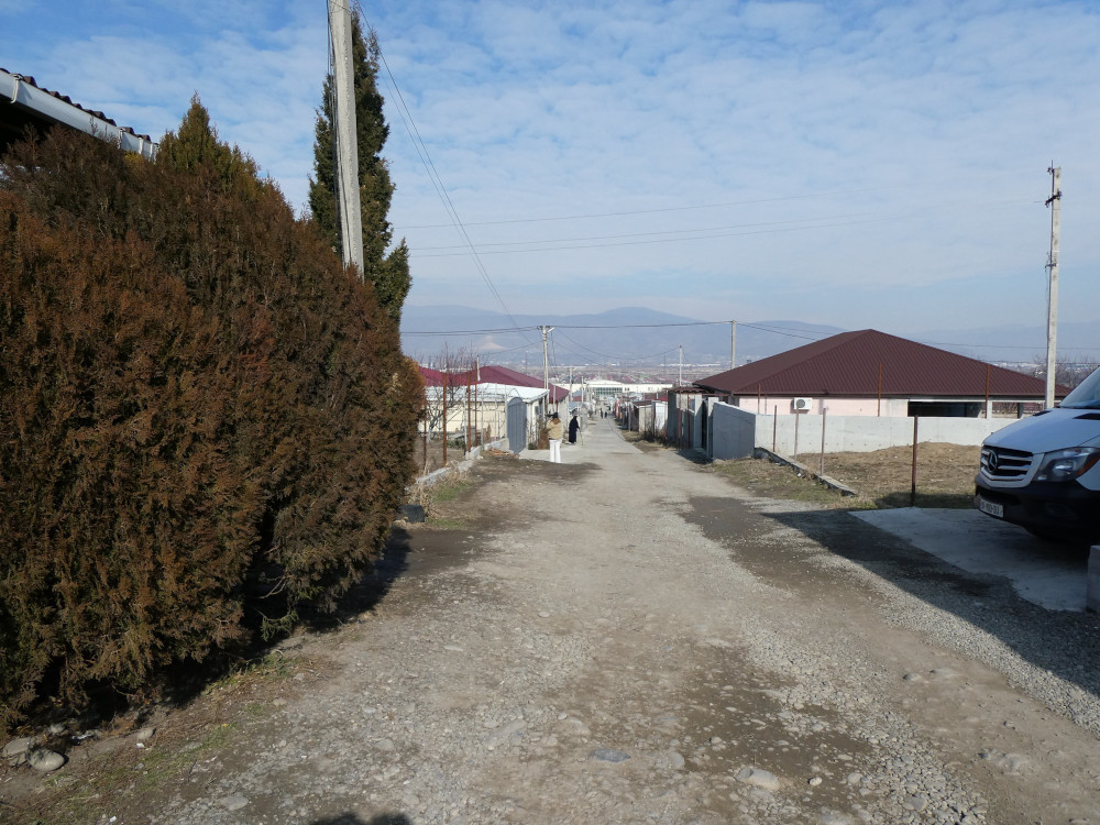 An unpaved street stretching into the distance towards some misty mountains. Down the side of the road are small white houses with red roofs and in the foreground is a large hedge.