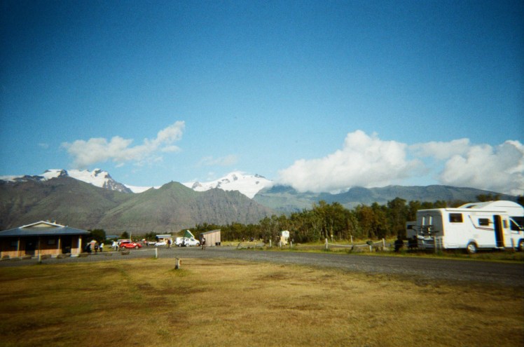 The view across Skaftafell campsite, featuring short yellow grass and a glacier on top of a wall of mountains in the distance.