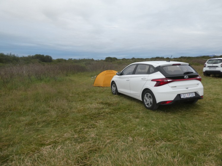 My little black tent half-hiding behind my white Hyundai i20 in a very basic field with some scrubby bushes.